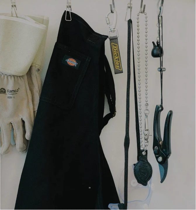 Black Dickies work apron with chest pocket, hanging on a wall beside leather gloves, a keychain and pruning shears