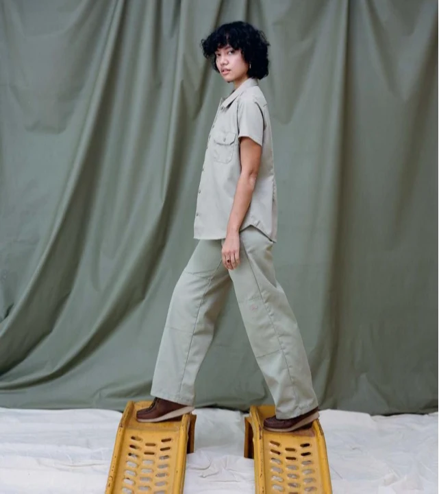 Woman in a sage Dickies short-sleeve work shirt and trousers stepping across two yellow car ramps