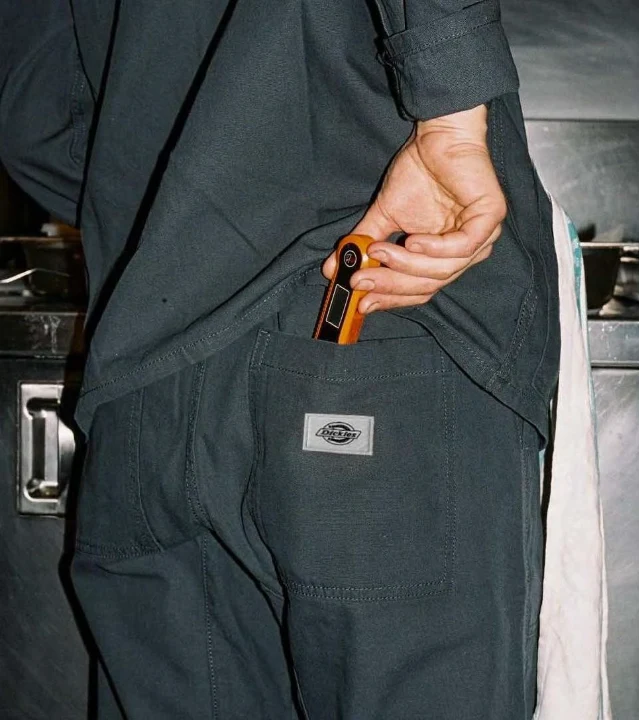 Hand reaching into the back pocket of charcoal Dickies work trousers, a Dickies label on the pocket, in a kitchen
