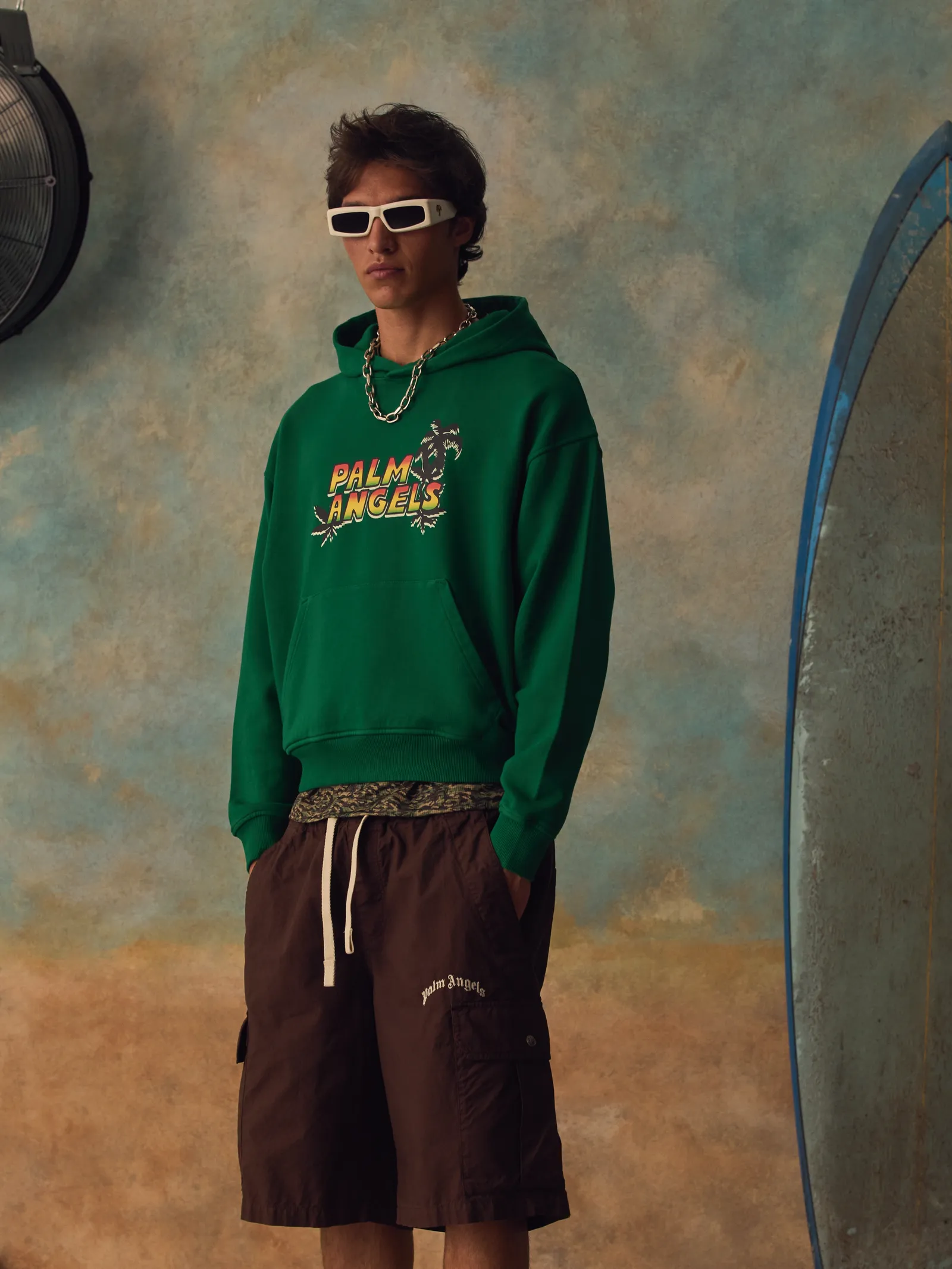 Model in white sunglasses, a green Palm Angels logo hoodie and brown cargo shorts, standing beside a surfboard