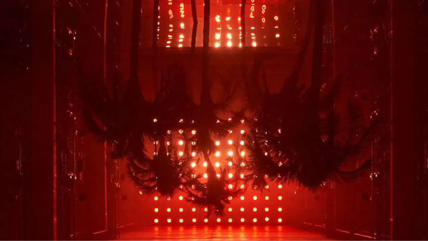 Red-lit runway set with dark hanging foliage and rows of glowing lights for a Palm Angels show