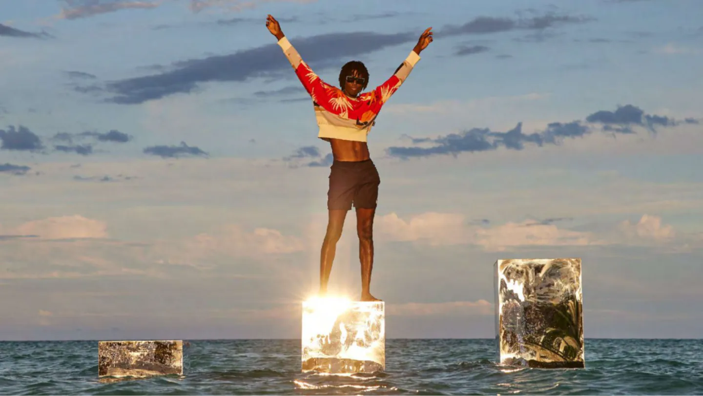 Model in a red palm-print sweater with arms raised, standing on a glowing glass block in the sea, Palm Angels
