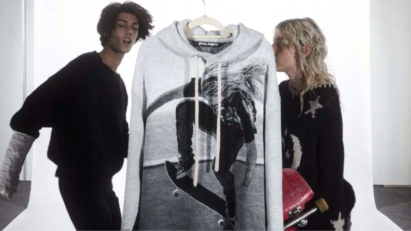 Two models flank a grey Palm Angels knit hoodie with a skateboarder print; one holds a red skateboard