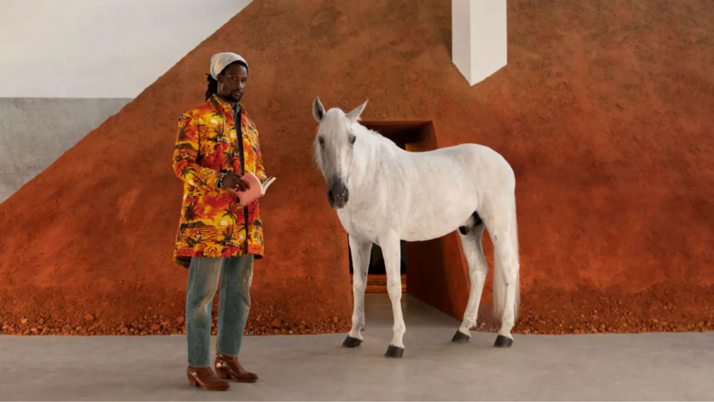 Model in a tropical sunset-print Palm Angels coat holding a book beside a white horse against a rust-red wall