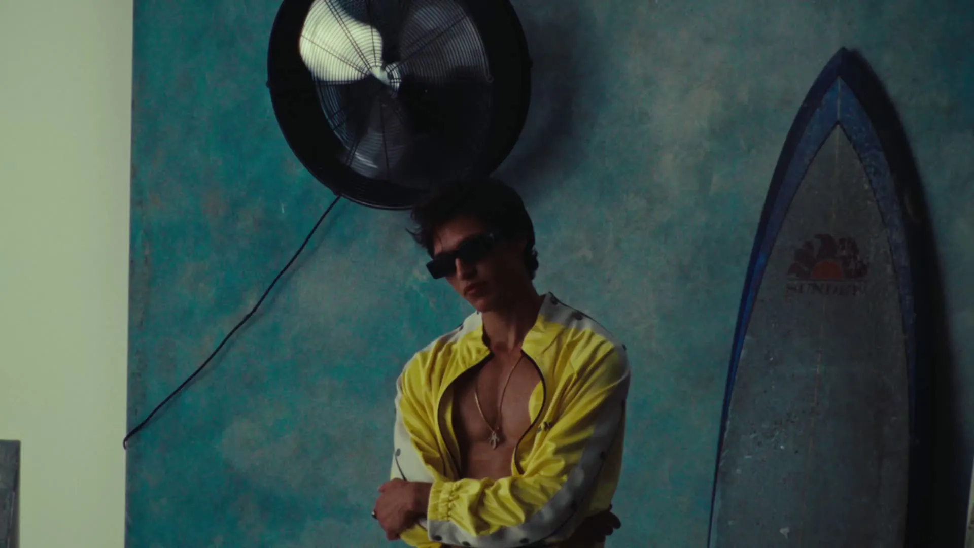 Model in a yellow track jacket and black sunglasses beside a surfboard in a Palm Angels campaign